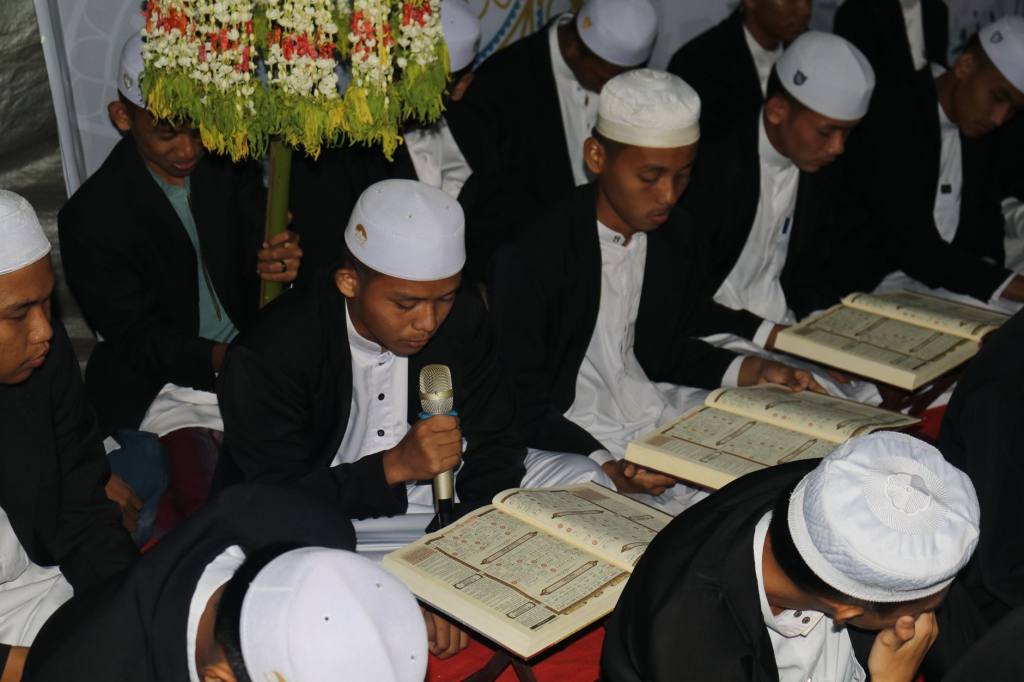 Men in traditional attire reading religious texts during a prayer gathering indoors.