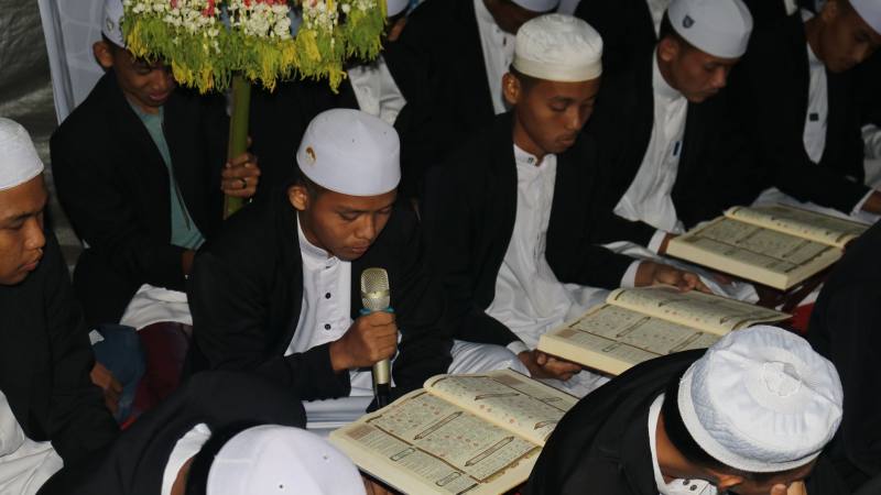 Men in traditional attire reading religious texts during a prayer gathering indoors.