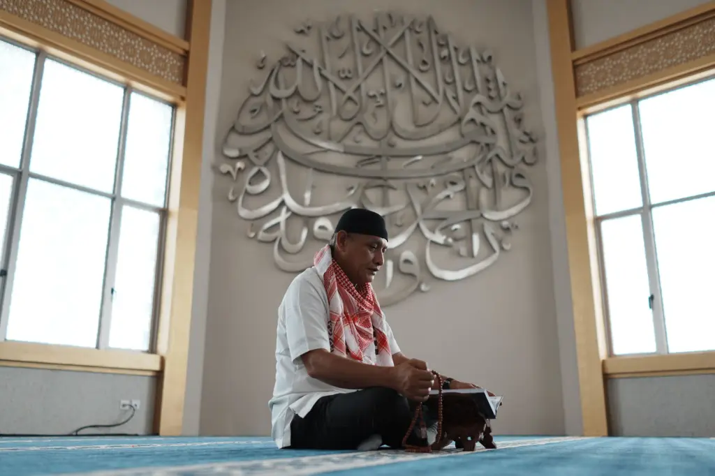 A man in traditional attire reading Quran inside a mosque in Indonesia, adorned with intricate calligraphy.