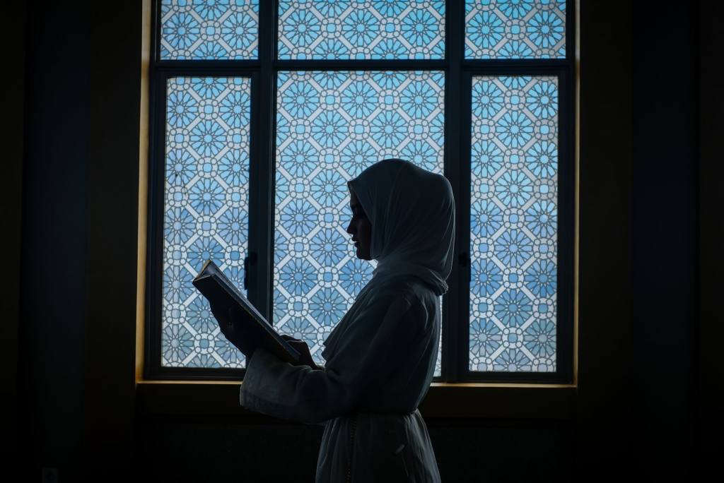 A Muslim woman reads the Quran by a decorative window, capturing a serene and spiritual moment.