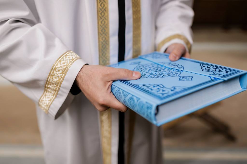 Close-up of a man holding a blue Quran, dressed in a traditional robe, signifying Muslim culture and religion.