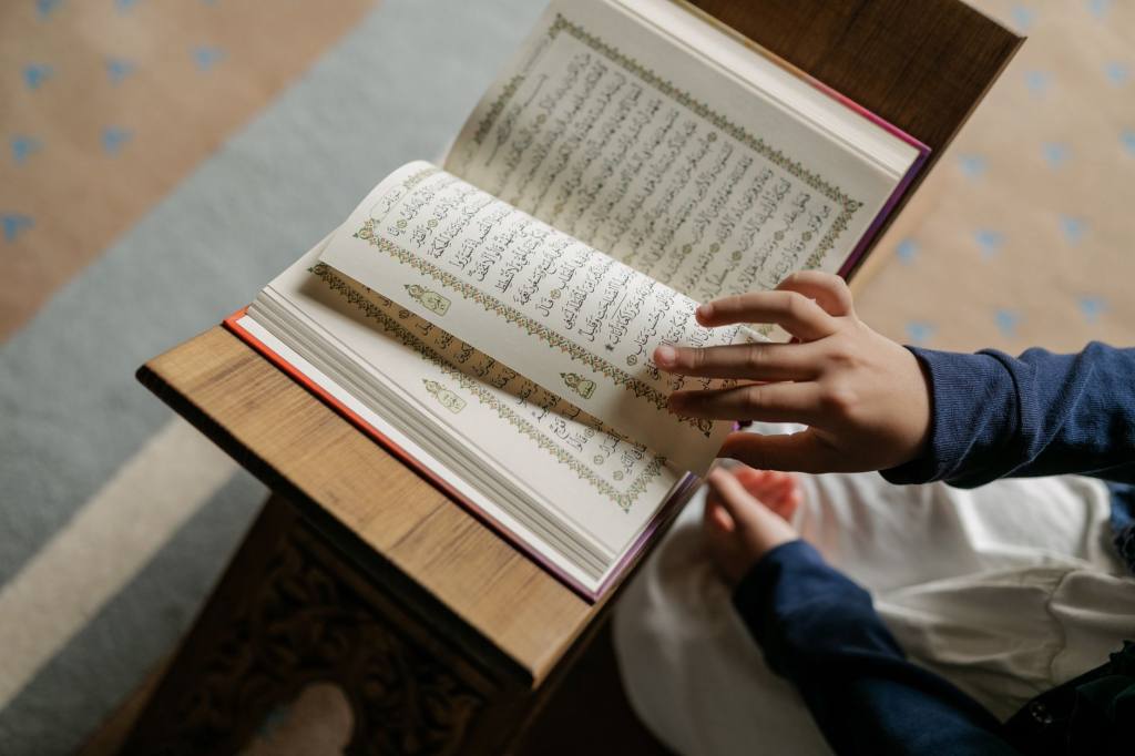 Child wearing hijab reading the Quran indoors, symbolizing Islamic culture.