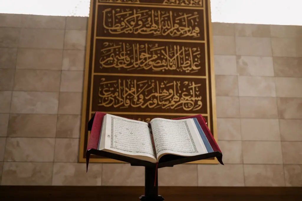An open Quran on a wooden stand in front of ornate Islamic calligraphy inside a mosque.