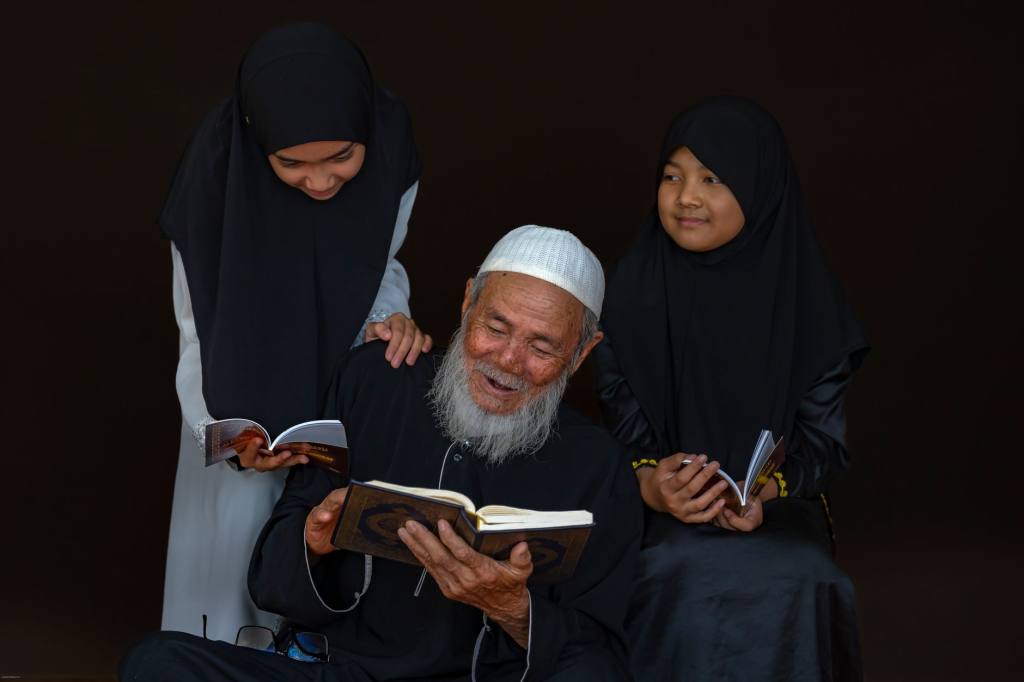 An elderly man and two young girls joyfully read books together, emphasizing family and cultural tradition.